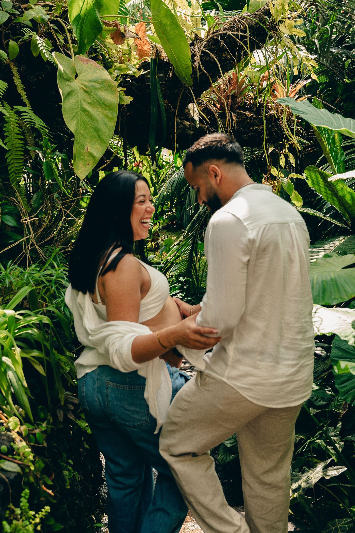 Happy pregnant couple dancing together surrounded by lush tropical plants in a botanical greenhouse.