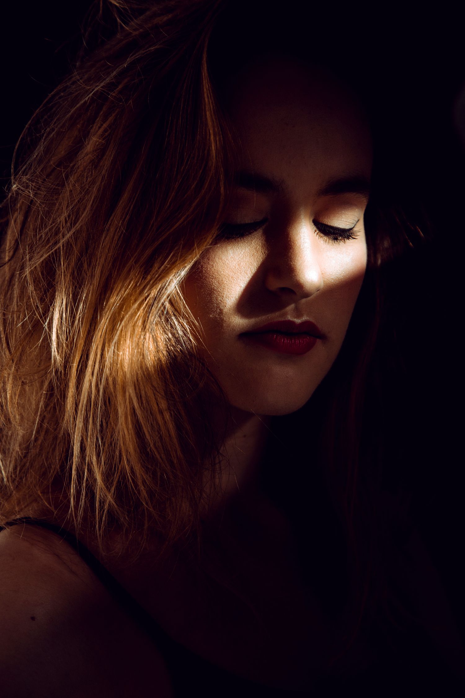 Moody low-key portrait of a woman with closed eyes and red lips illuminated by warm golden light.