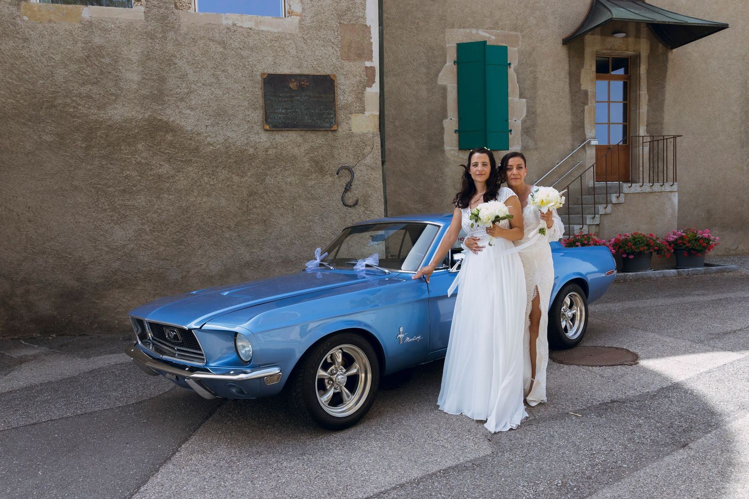 Two brides in white dresses posing beside a classic blue Ford Mustang convertible at their wedding.