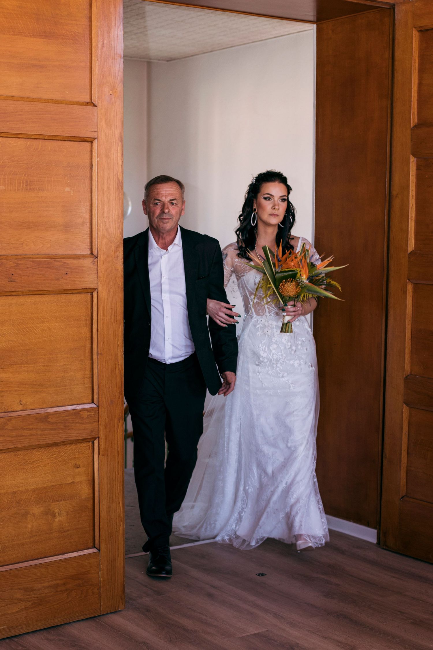 Bride in lace wedding gown holding tropical bouquet walks through wooden doors escorted by older man.