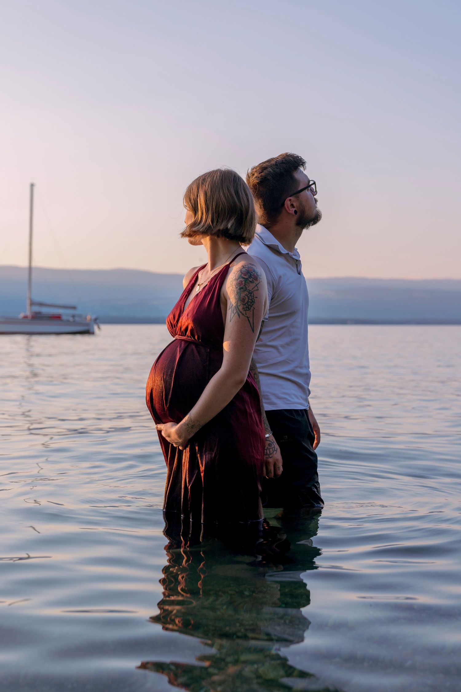 Pregnant woman and partner standing back-to-back in a lake at dusk with a sailboat nearby.