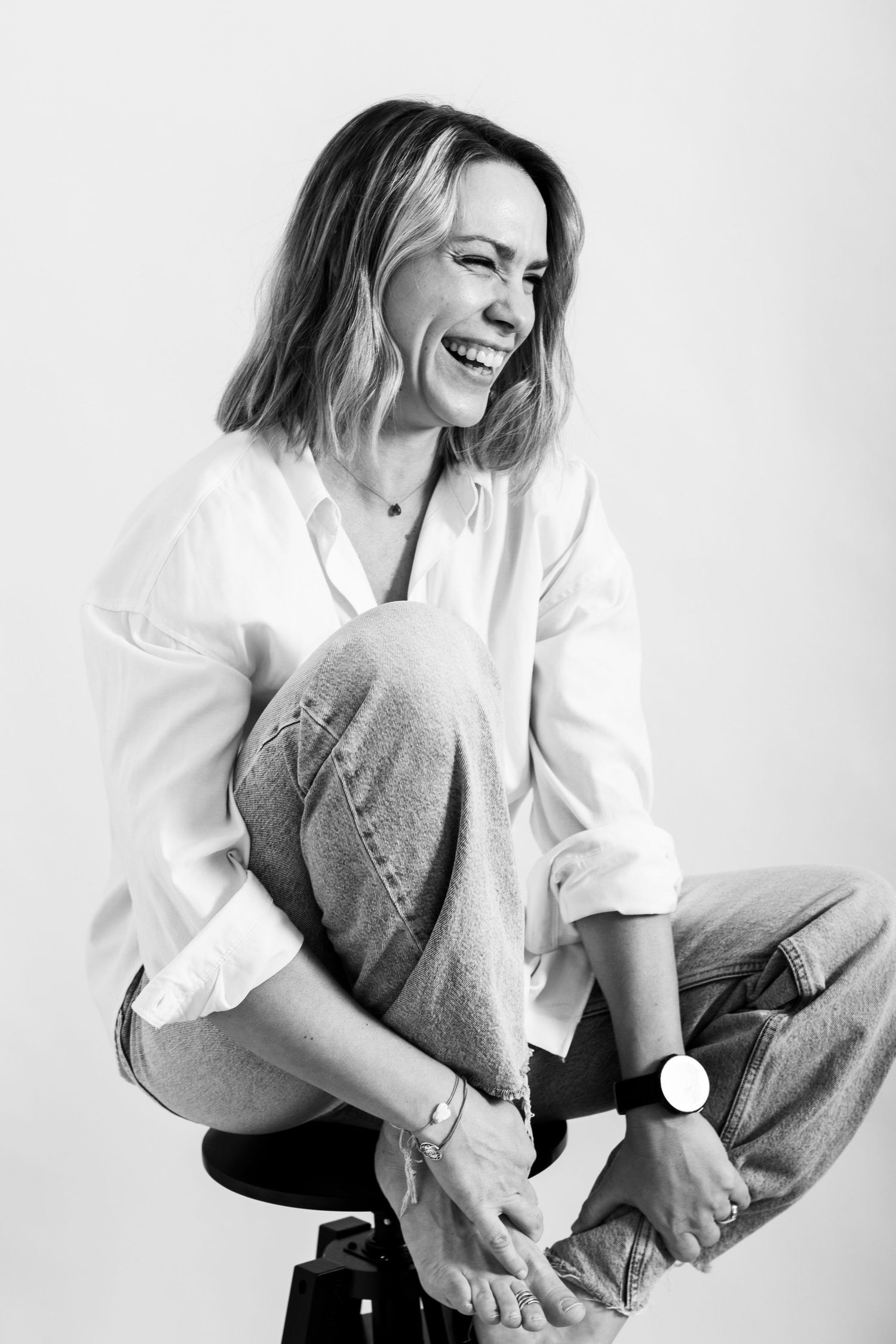 Laughing woman sitting on stool in white shirt and jeans, black and white portrait photo.