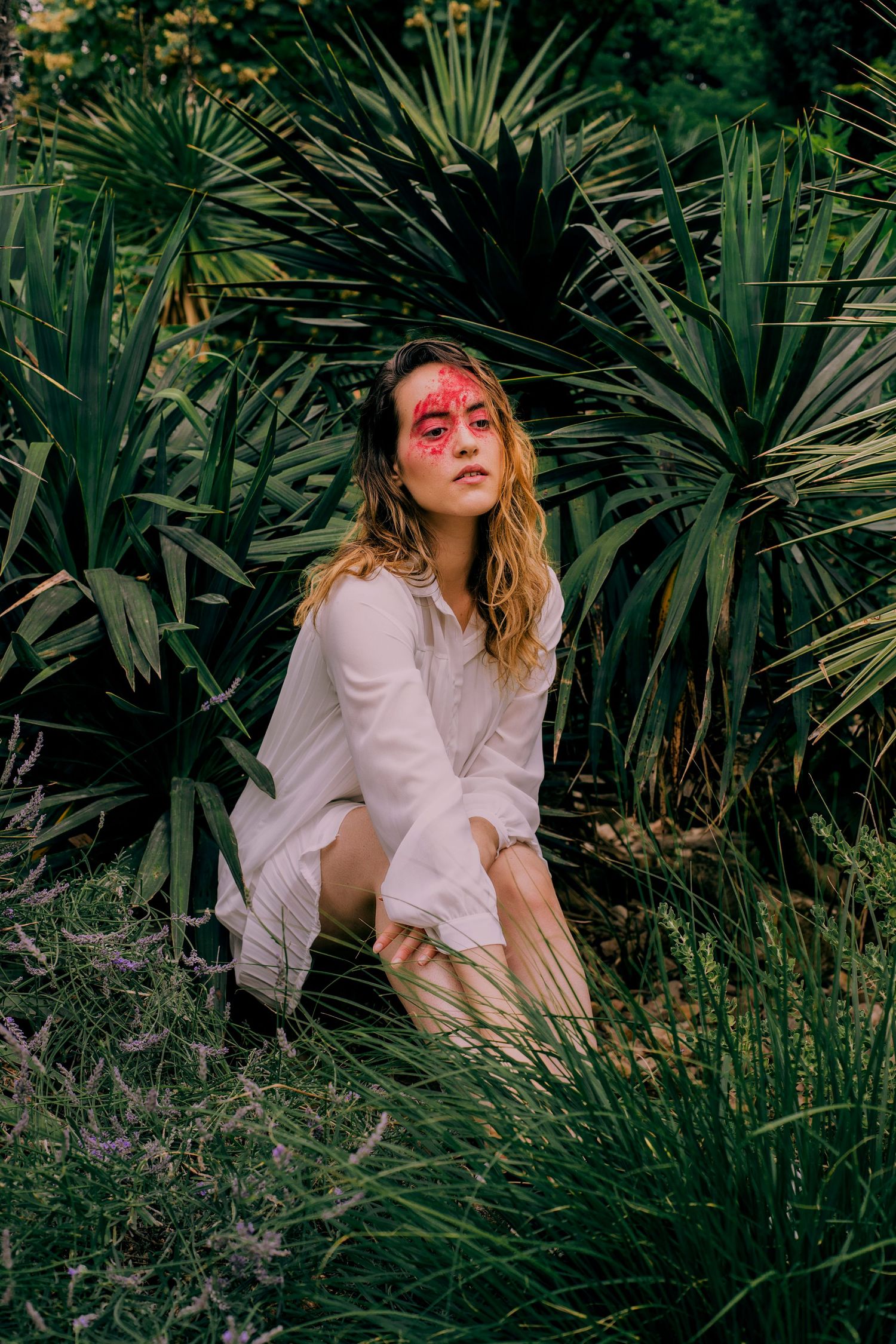 Woman with red face art in white shirt crouching among tropical garden plants.