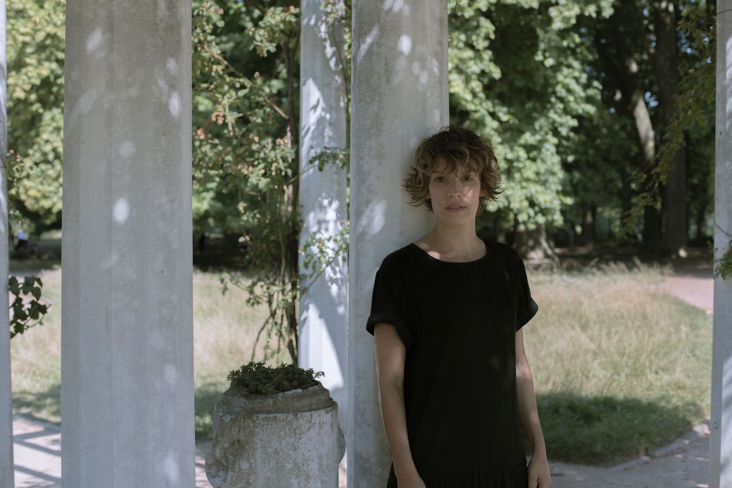 Young woman with curly hair in black top standing between white columns in a sunlit park.