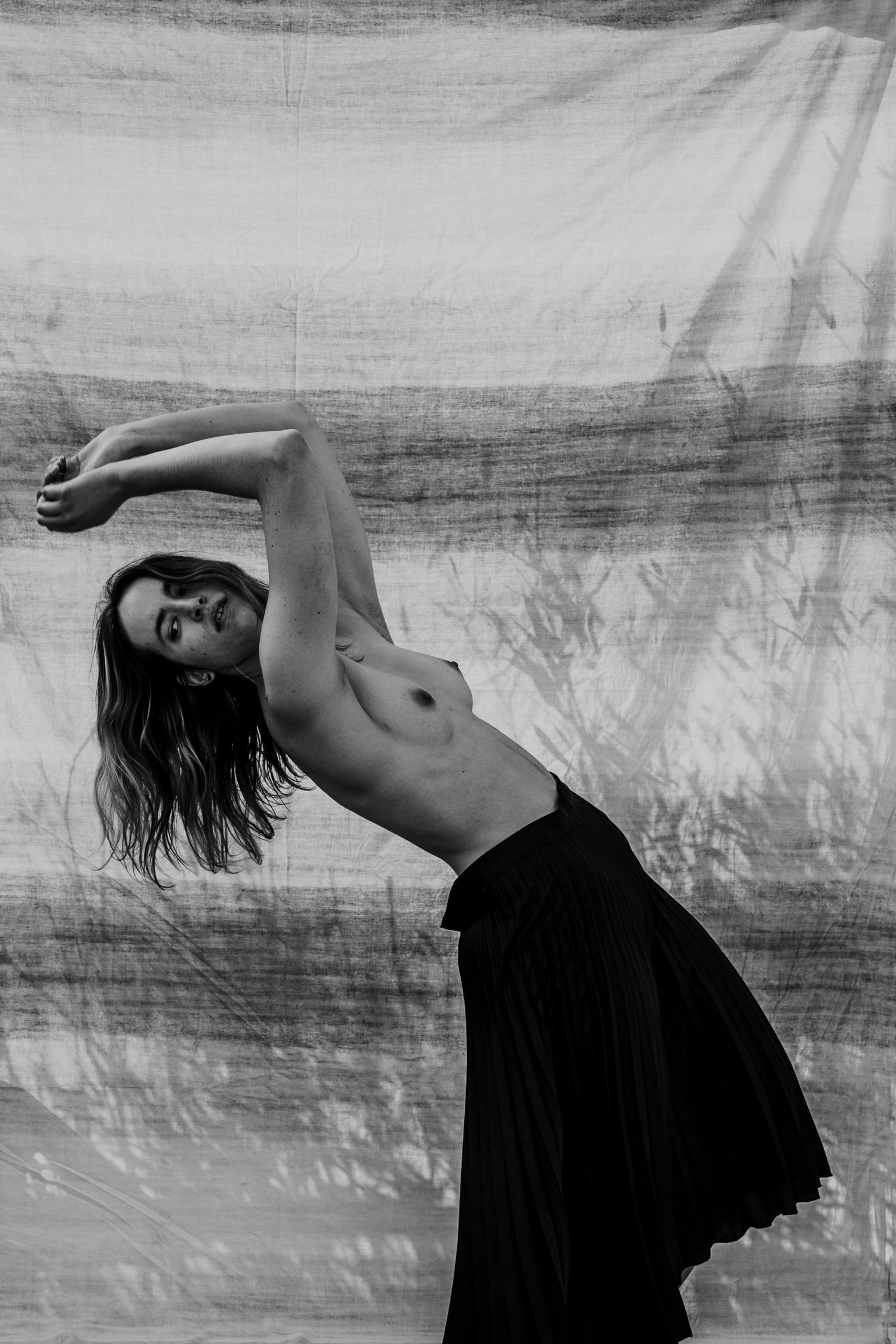 Dynamic black and white portrait of a topless woman in black skirt, leaning back with arms raised, fabric backdrop.