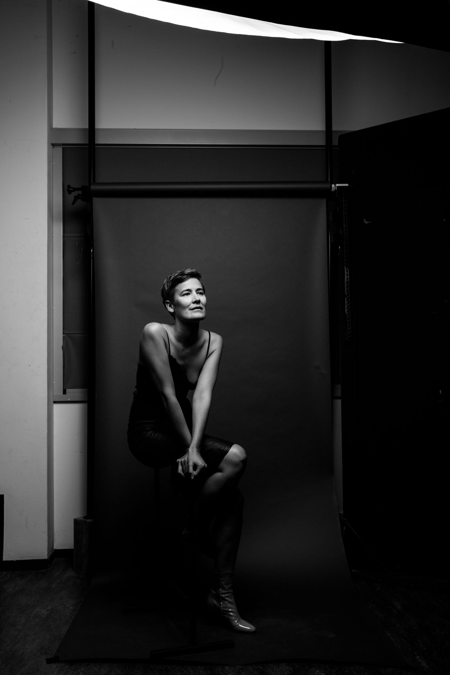 Woman seated on stool in dark studio with dramatic overhead lighting in artistic black and white portrait.