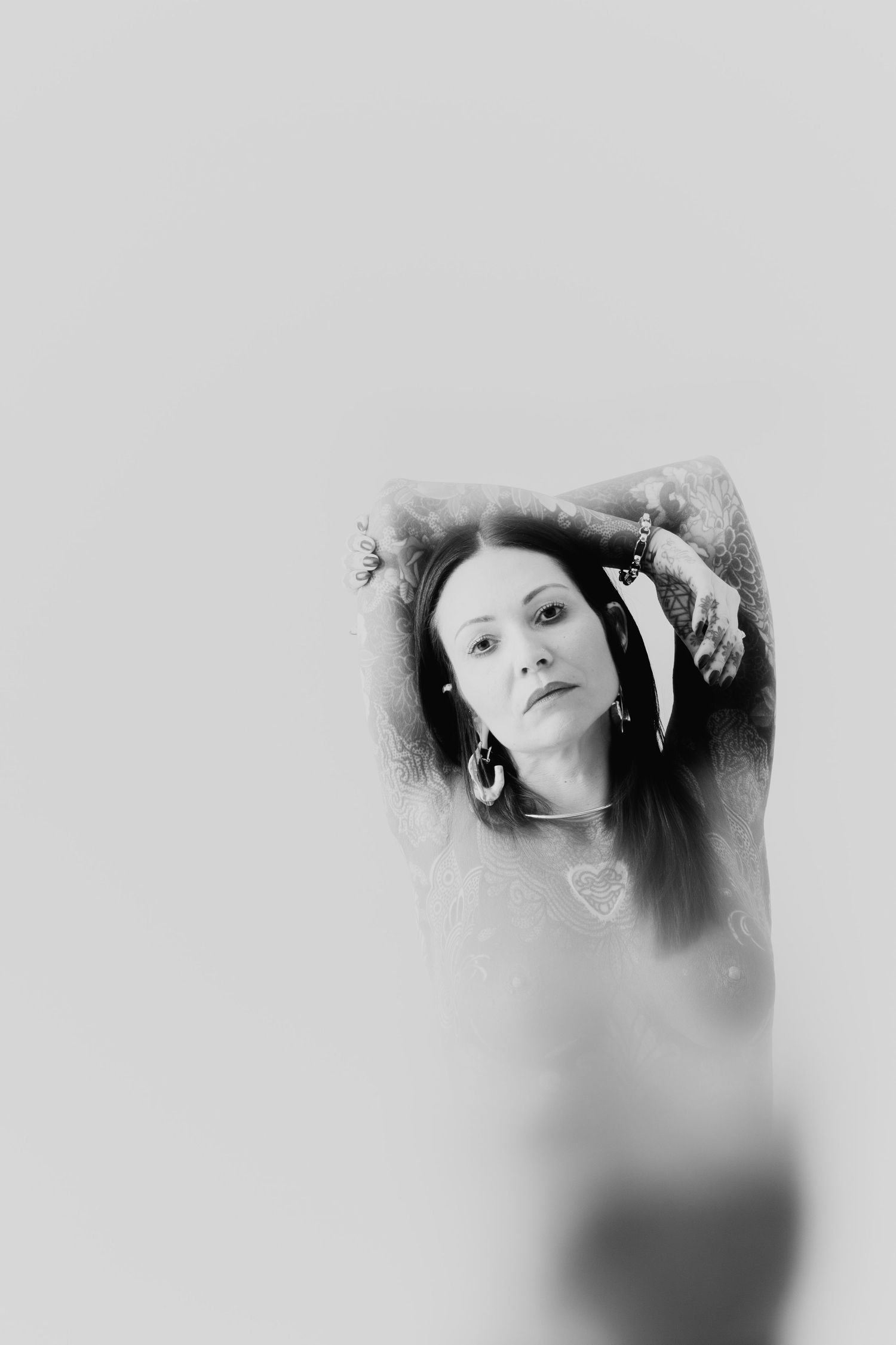 Black and white portrait of tattooed woman with arms raised above head gazing at camera.