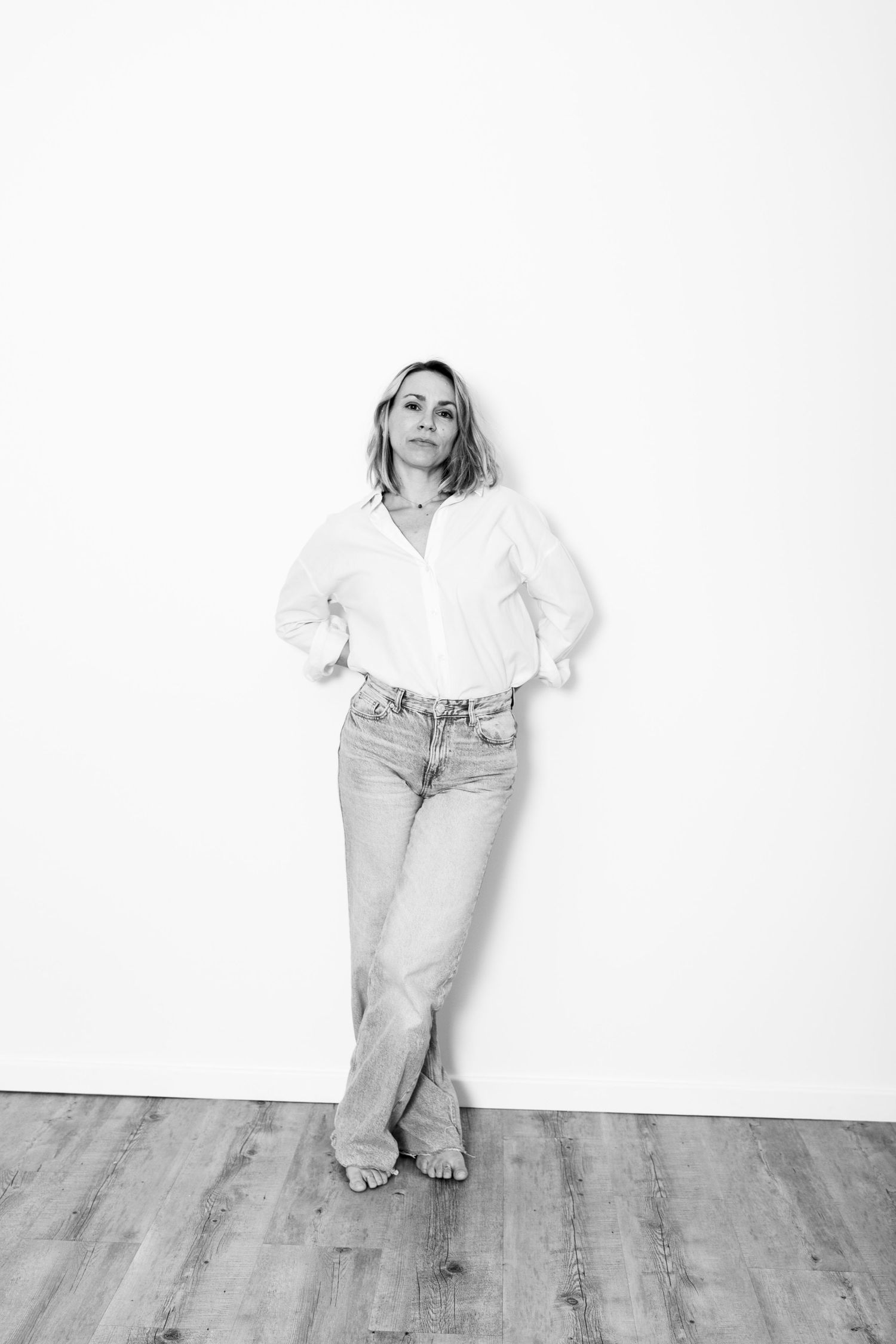 Woman in white shirt and jeans standing against white wall, black and white portrait.