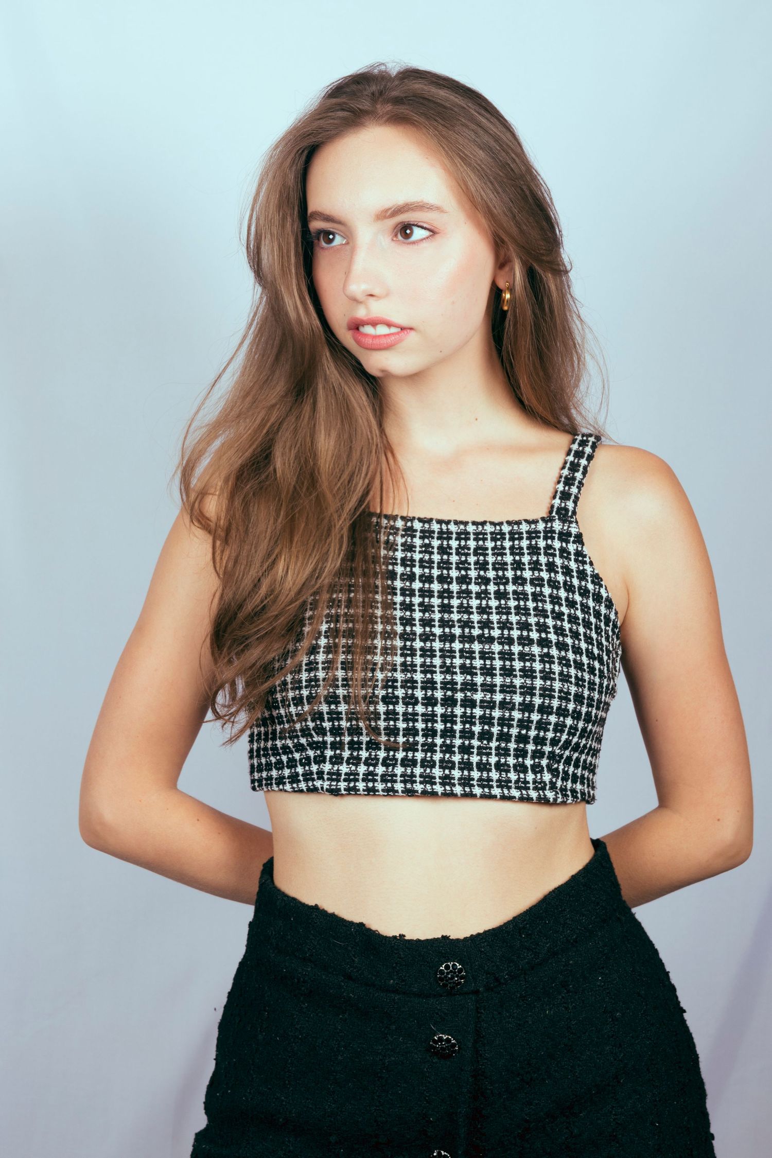 Young woman with long brown hair in checkered crop top and black buttoned skirt posing against light blue background.