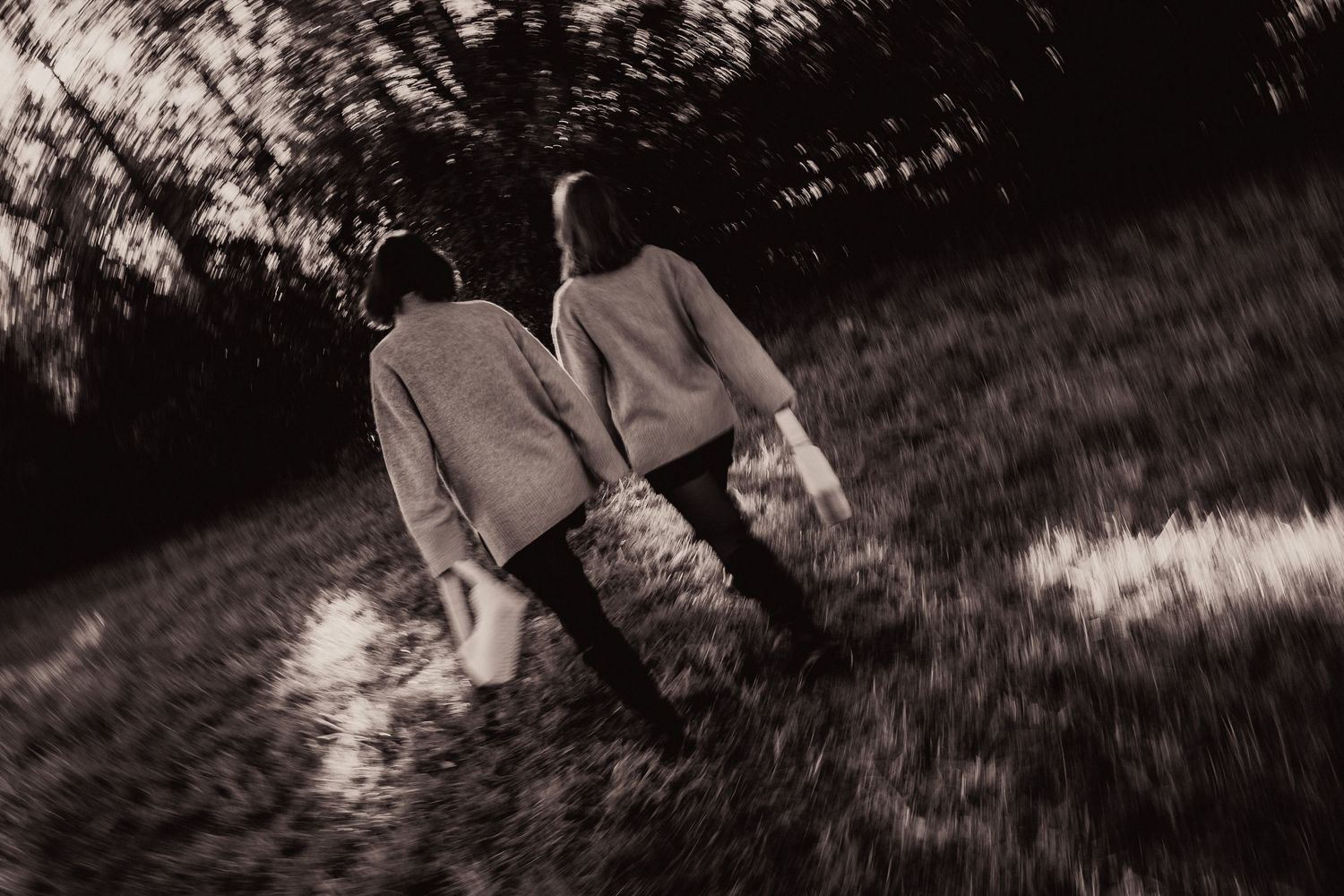 Black and white rear view of two women in matching sweaters walking along a sunlit wooded path.