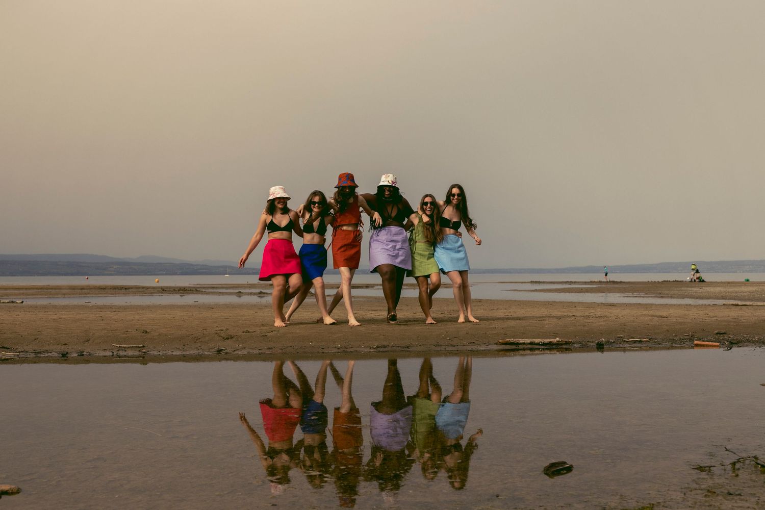 Group of six women in colorful outfits walking along a beach at sunset with reflections in water.