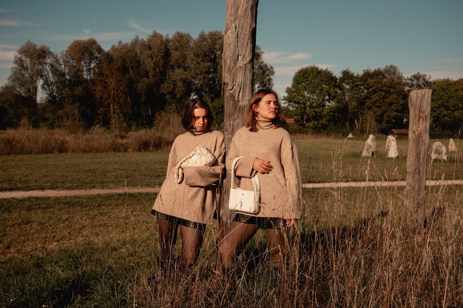 Two women in matching beige oversized sweaters holding white bags, standing by a fence in golden hour light.
