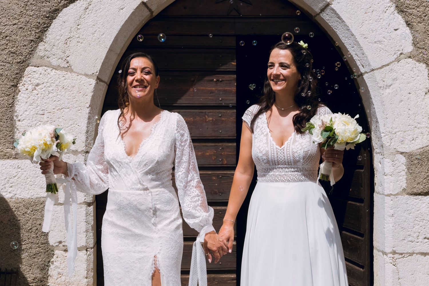 Two brides in white dresses holding bouquets and smiling as they exit through a stone archway.