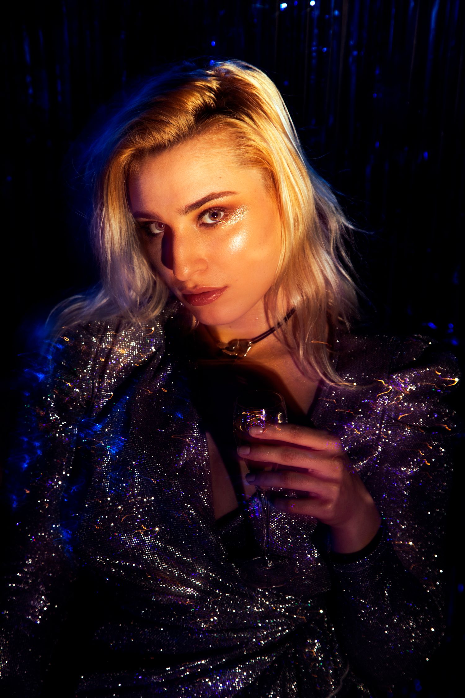 Blonde woman in a glittery jacket holding a drink under blue neon lights at a party.