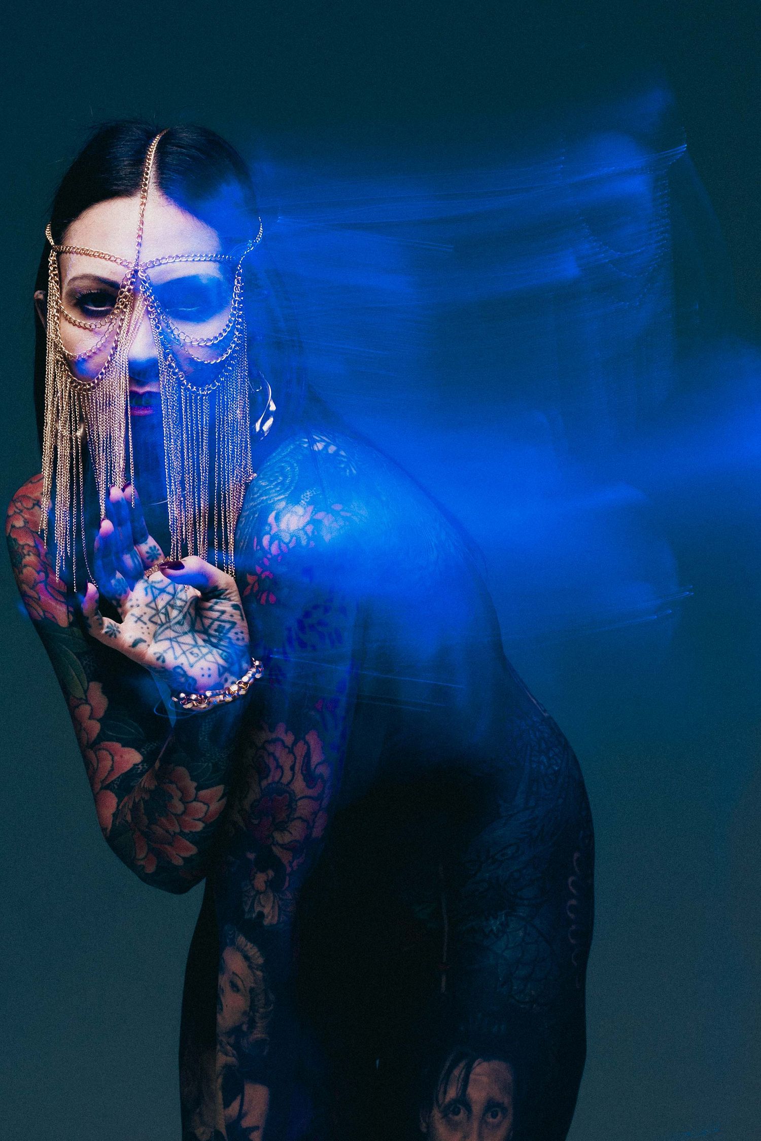 Tattooed woman holding decorative mask with blue light motion blur in dark studio portrait.
