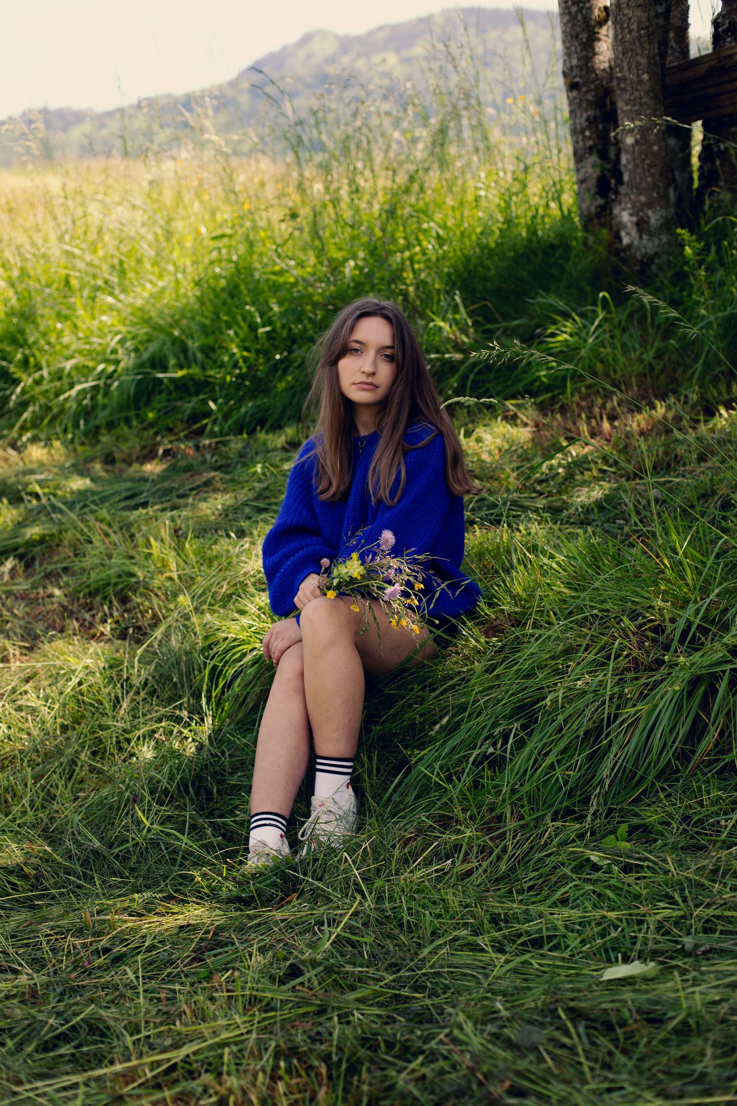 Young girl in blue hoodie sitting on grass holding wildflowers with mountains in background.