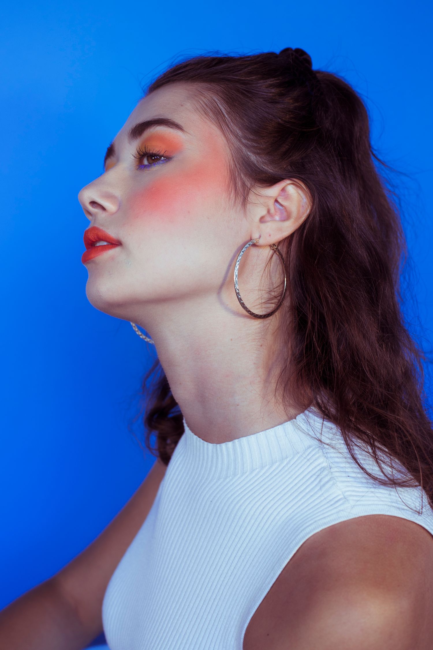 Profile of woman with vibrant orange blush and hoop earrings on blue.