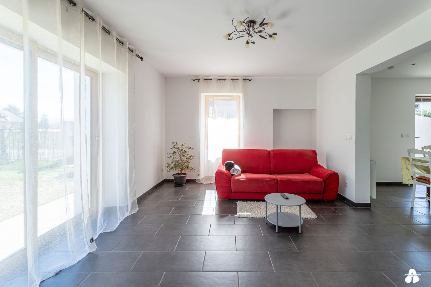 Bright living room with red sofa, dark tile floors, white curtains and a small round coffee table.