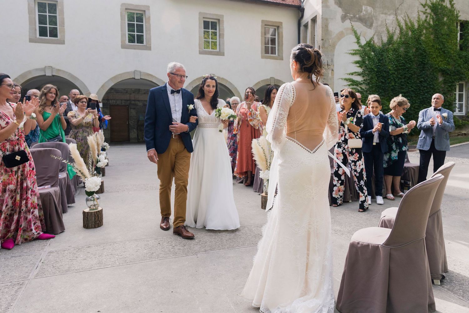 Two brides walk down the aisle at an outdoor wedding ceremony surrounded by applauding guests.