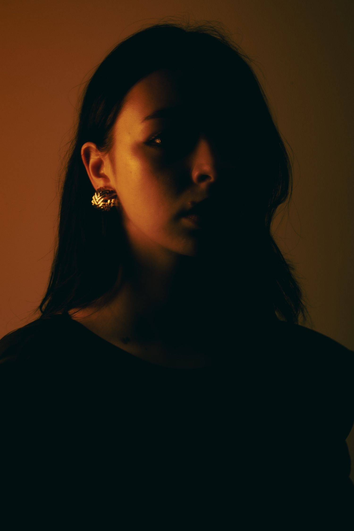 Dramatic low-key portrait of young woman wearing gold earring against warm amber background.