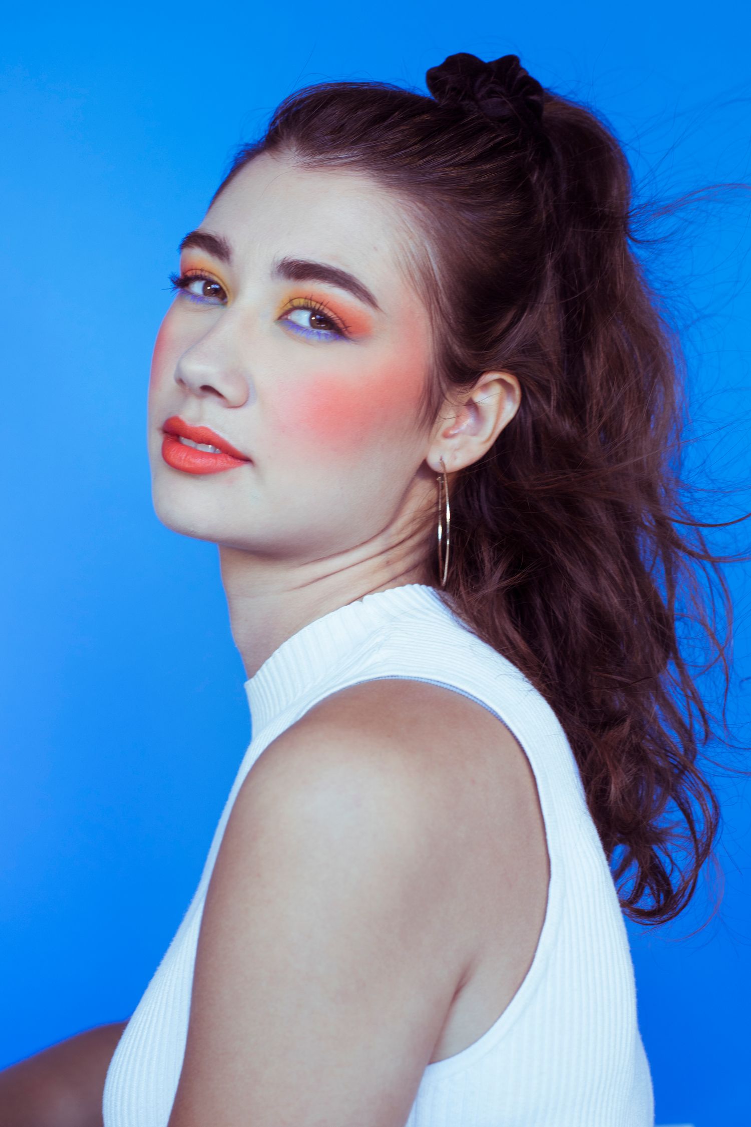 Brunette model with striking orange makeup and hoop earring in white turtleneck against blue backdrop.