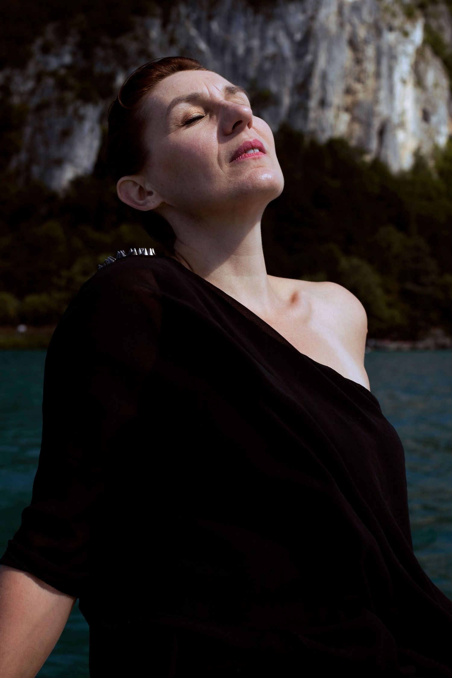 Woman in black off-shoulder top with eyes closed, basking in sunlight near turquoise water and rocky cliffs.
