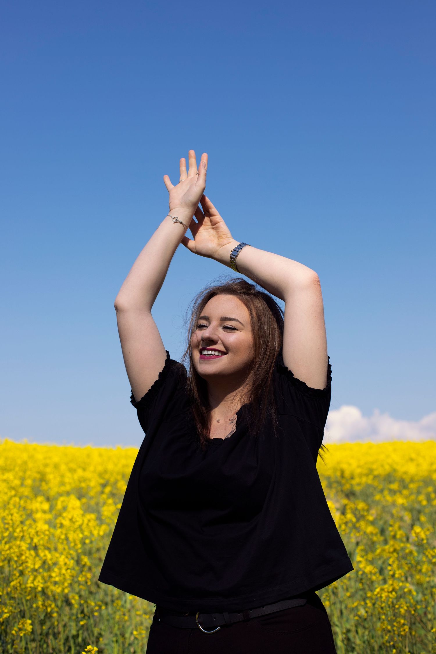 Happy young woman in black top raising hands above head in a vibrant yellow rapeseed field.
