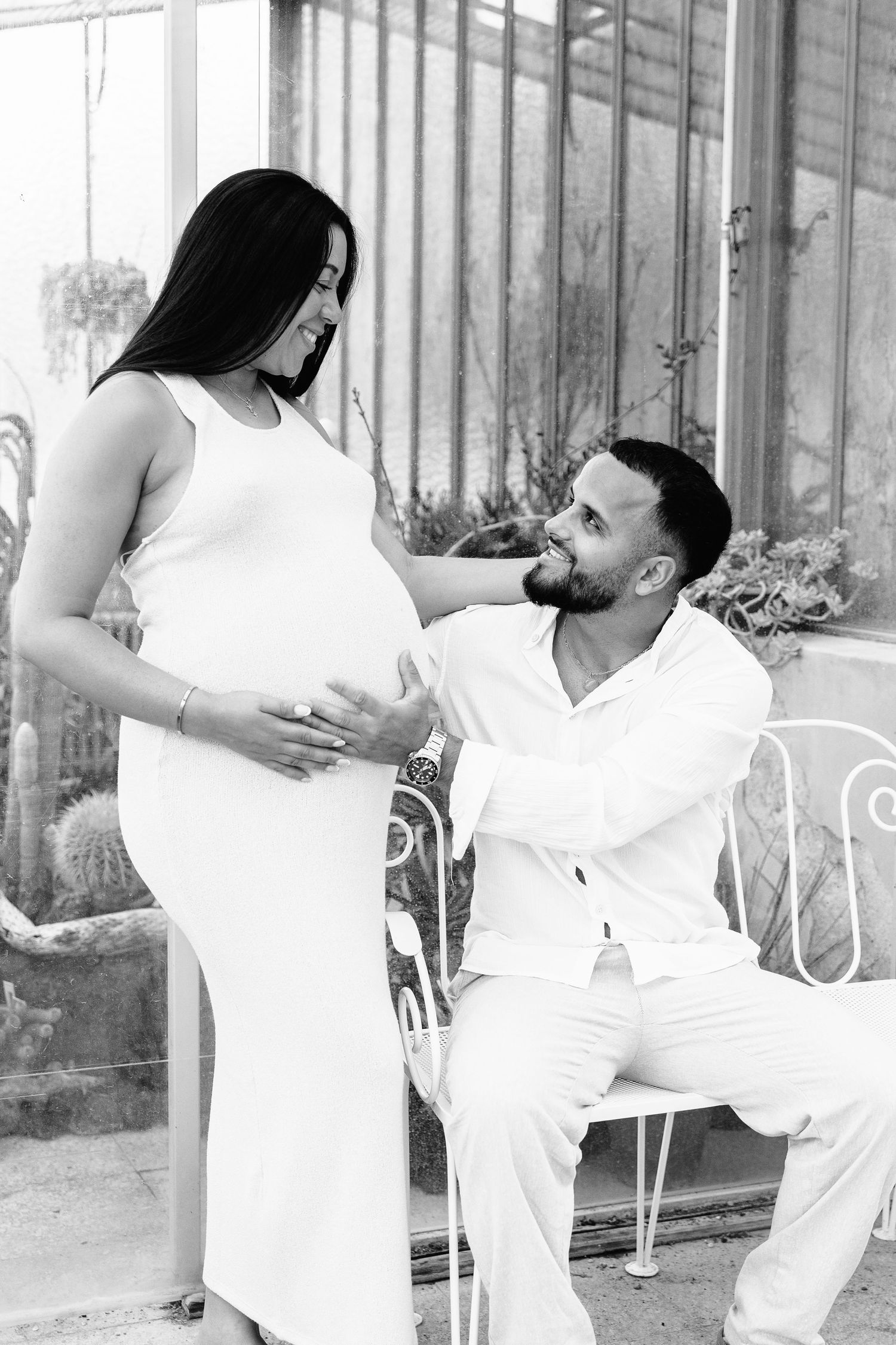 Black and white maternity photo of pregnant woman in white dress with seated man tenderly touching her belly.