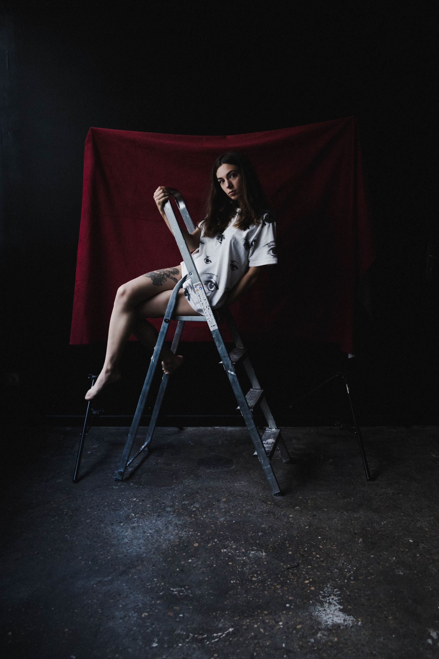 Young woman in white graphic tee sitting on metal ladder against dark background with red drape.