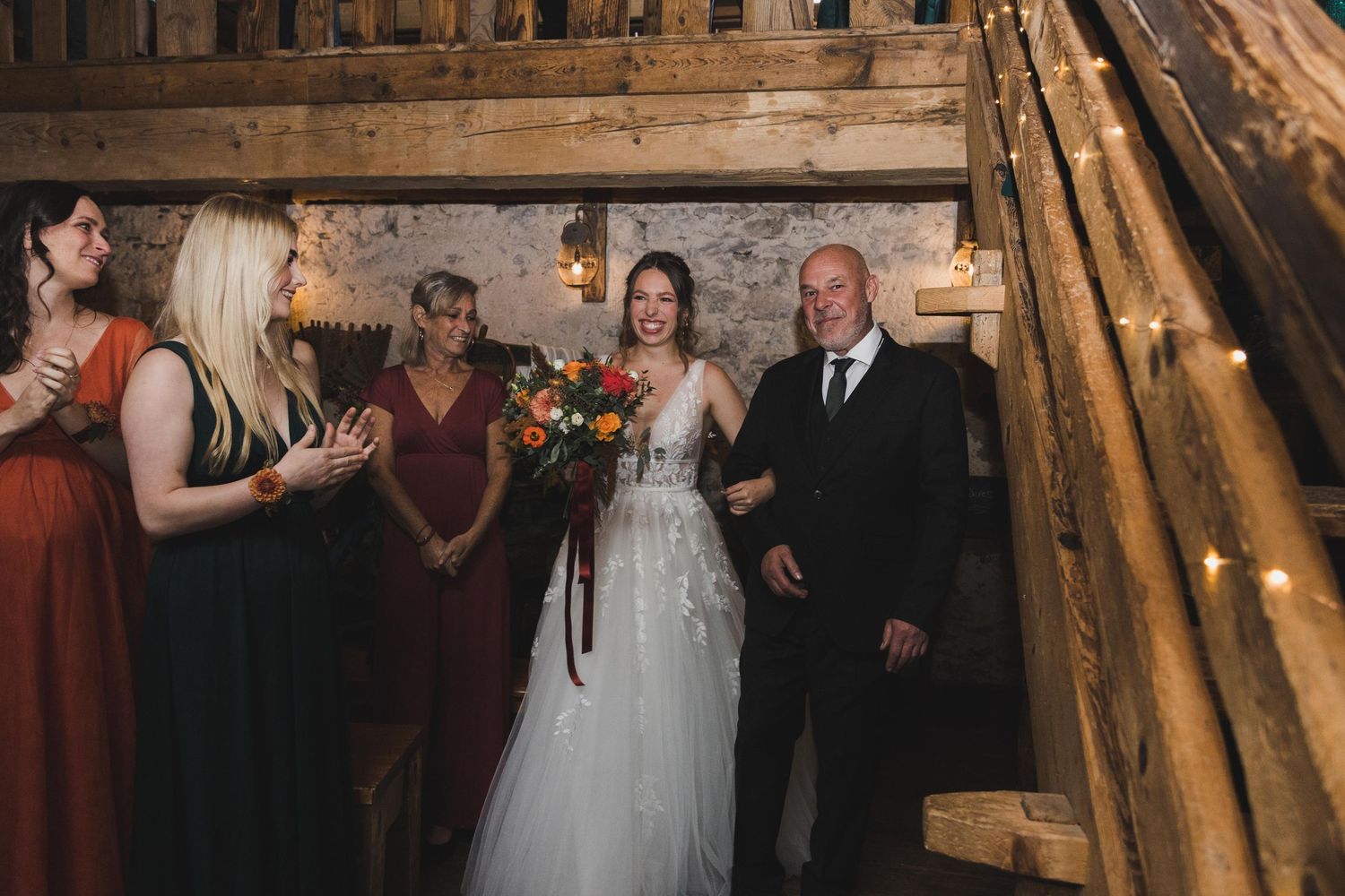 Bride in lace gown holds colorful bouquet with father and bridesmaids in green dresses.