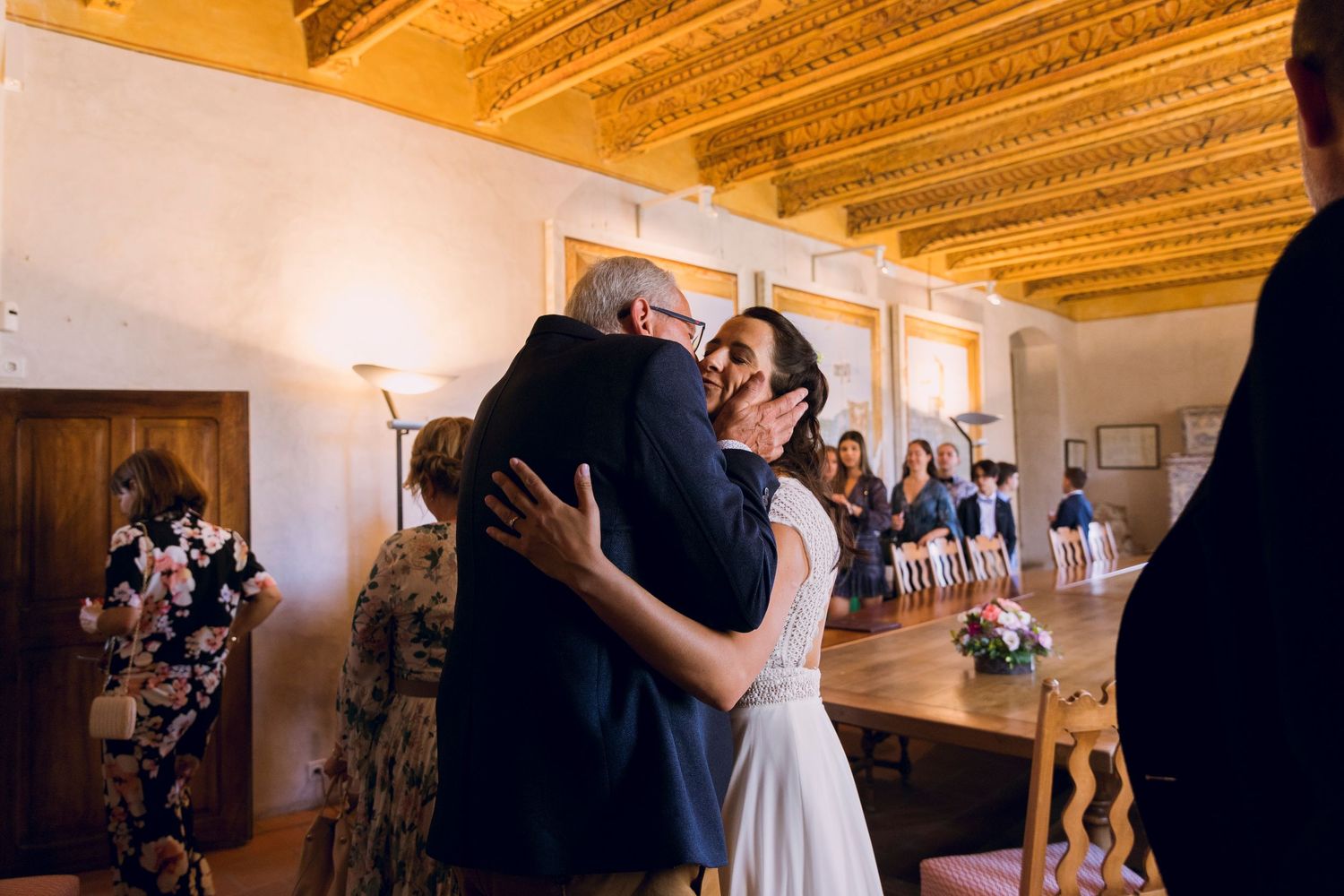 Bride in white dress sharing a warm embrace with an older guest inside a rustic wedding venue.