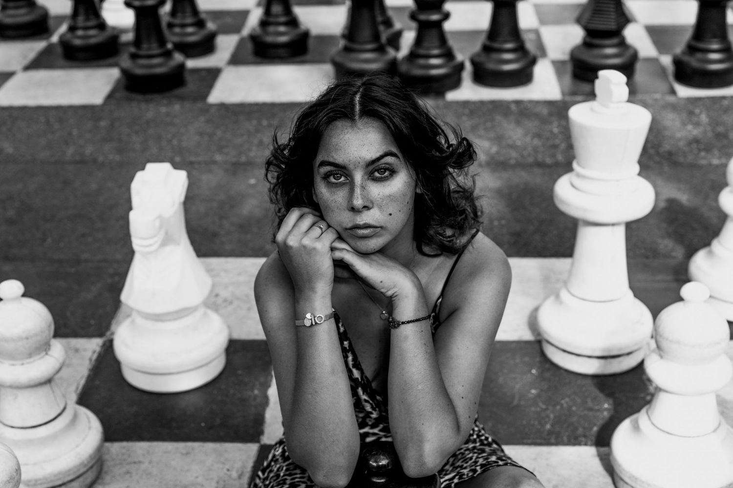 Black and white photo of woman sitting pensively on giant outdoor chess board with pieces.