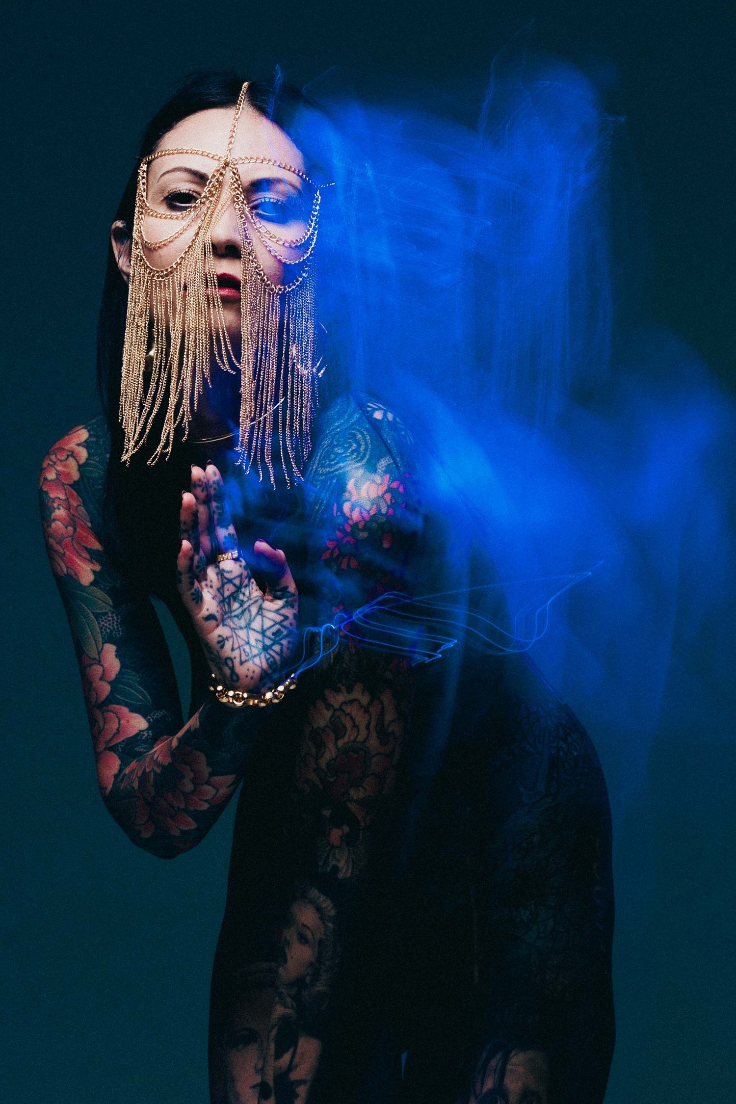 Tattooed woman in ornate chain mask with blue smoke swirling around her in dark studio.