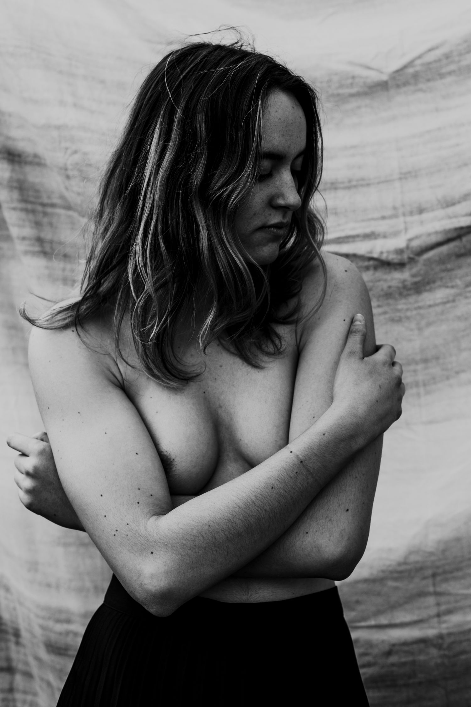 Artistic black and white portrait of a topless woman with eyes closed, arms crossed, against fabric backdrop.