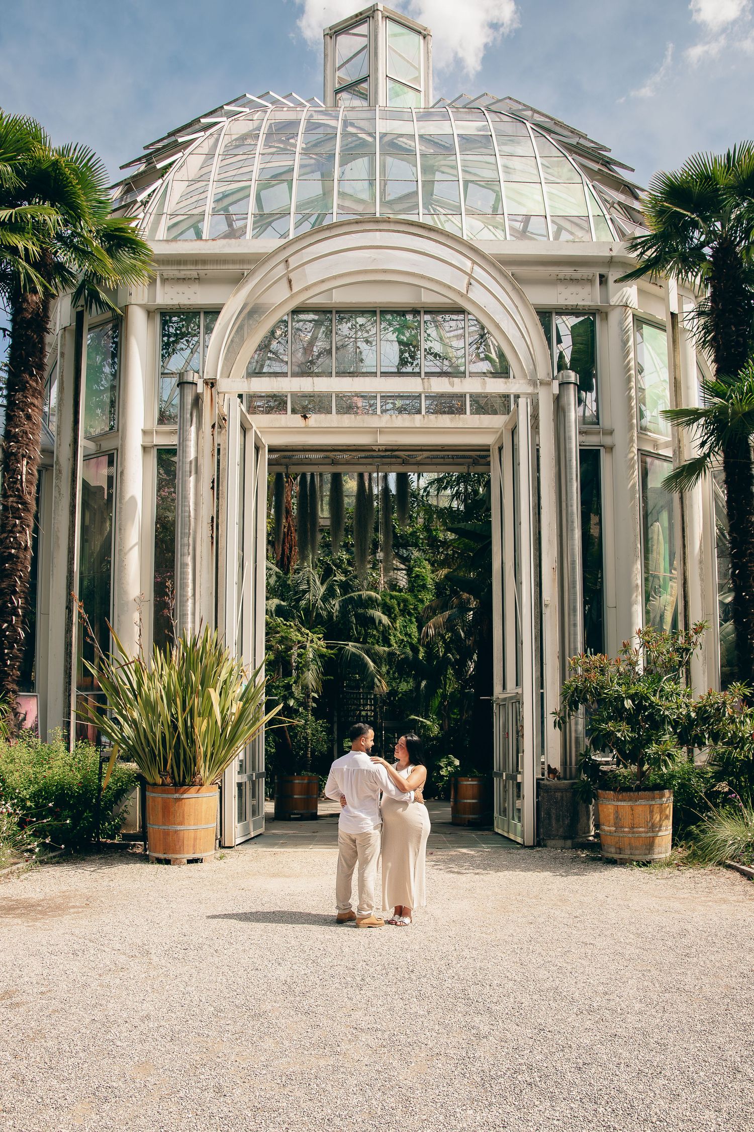 Couple in white outfits stroll toward the grand arched glass entrance of a botanical greenhouse.
