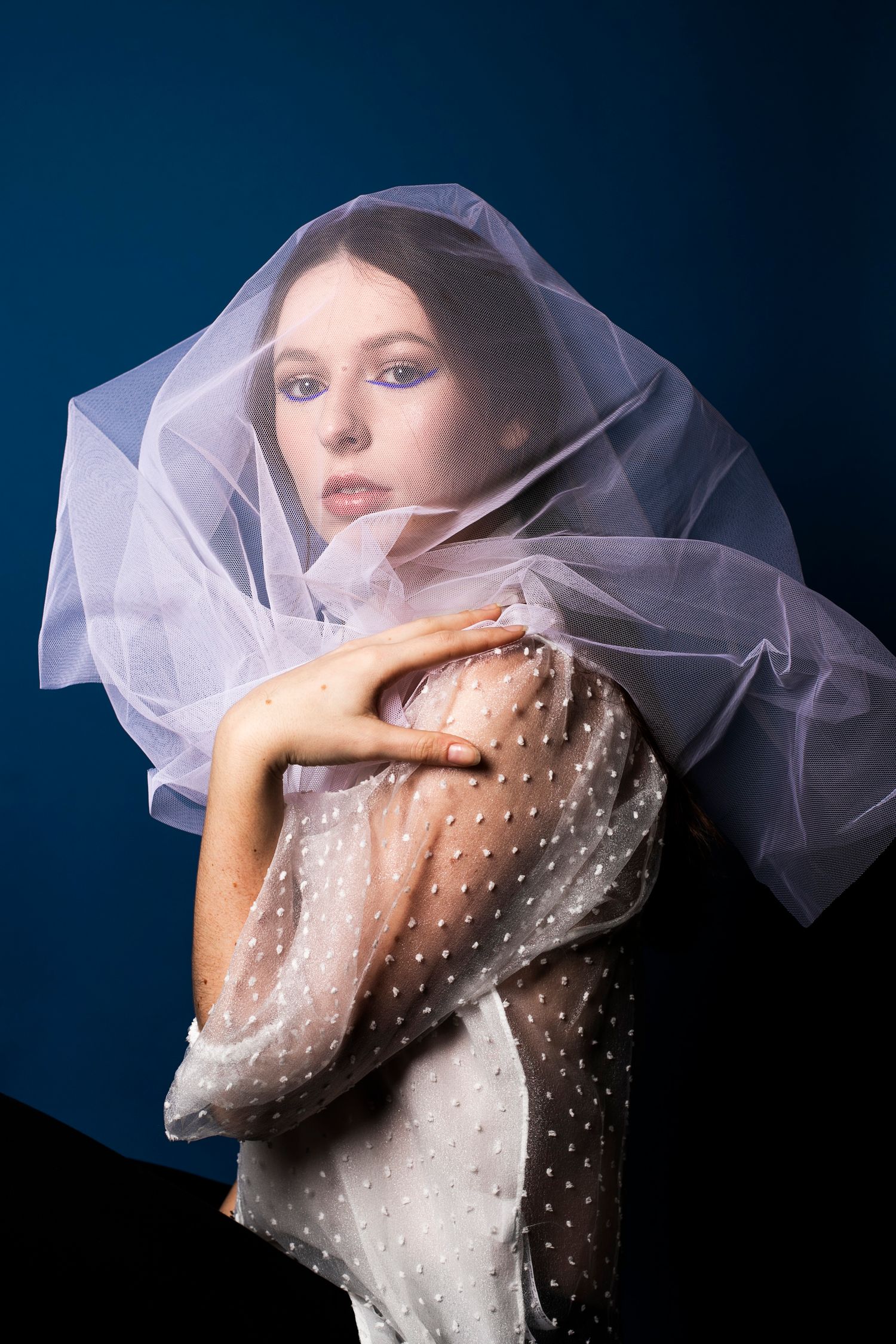 Elegant woman wrapped in a sheer white polka-dot fabric and veil against a deep blue background.