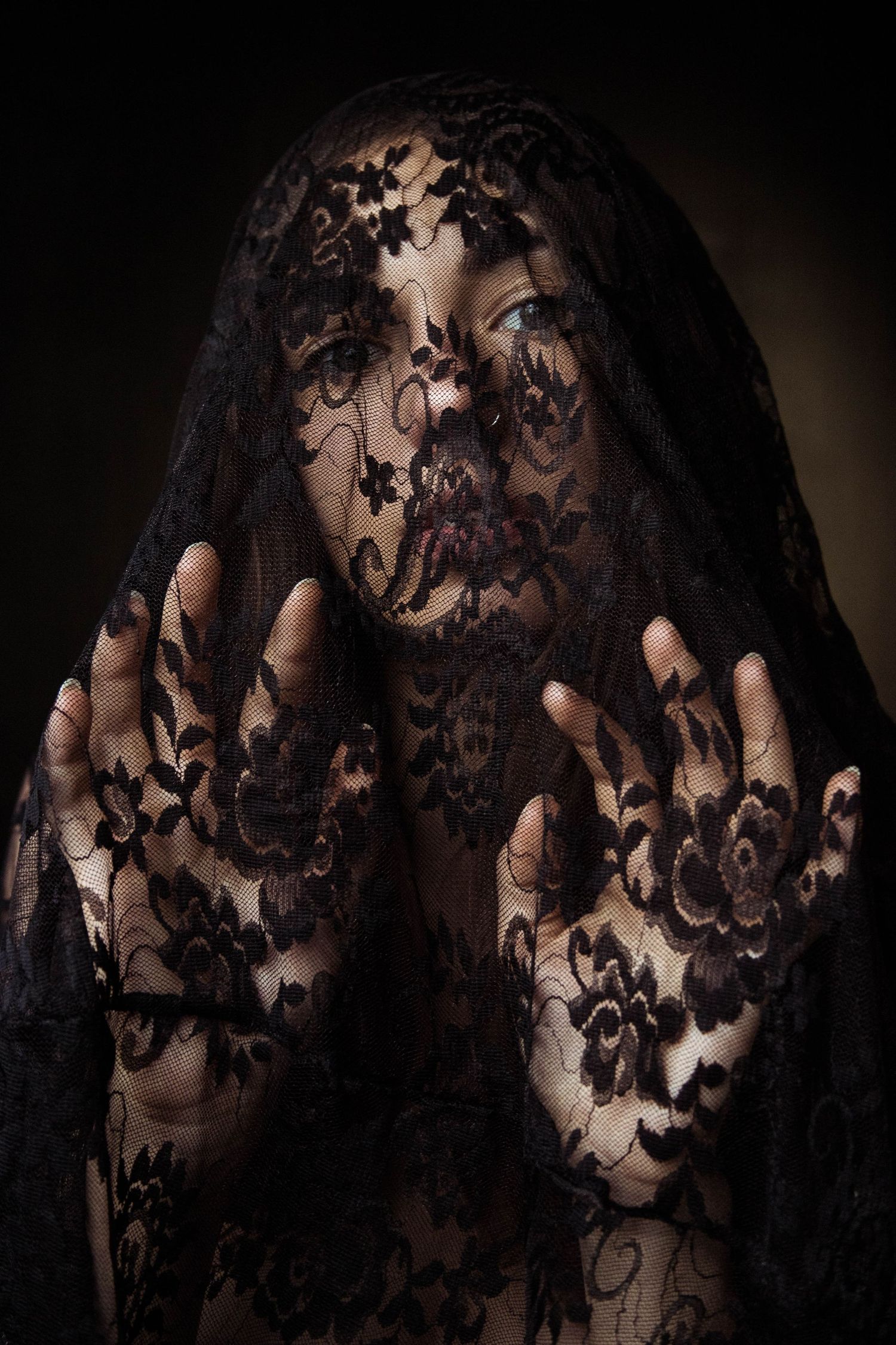 Woman covered in black floral lace veil with hands raised in prayer pose against dark background.