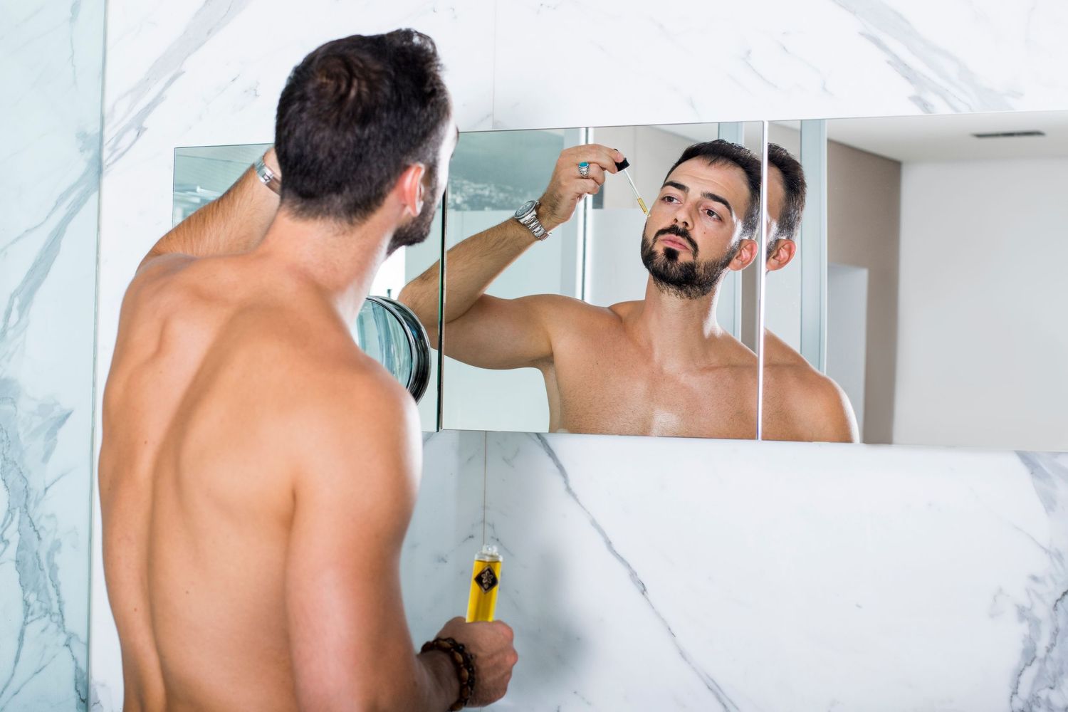 Shirtless bearded man applying product while looking in marble bathroom mirror.
