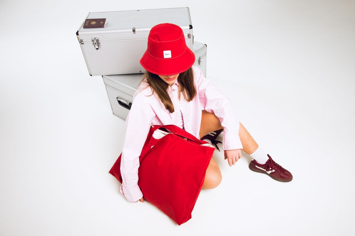 Model in red bucket hat sits on floor holding red canvas tote bag next to silver trunks.