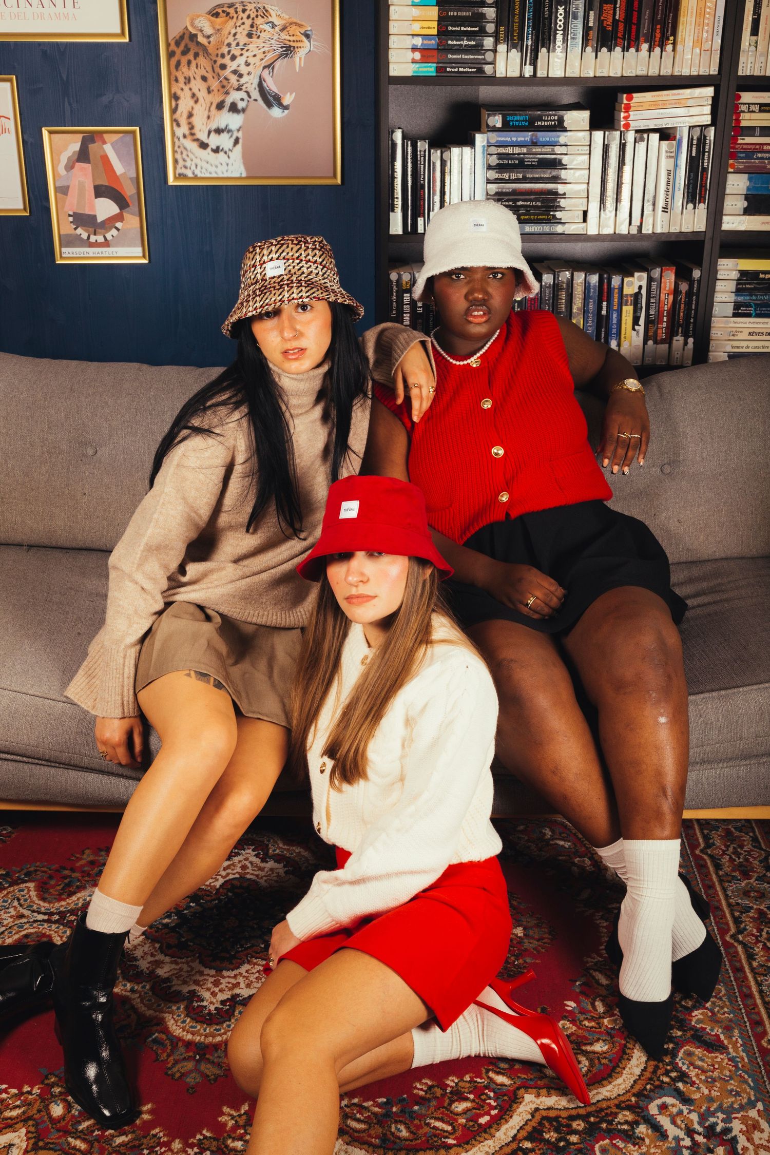 Three fashionable women in bucket hats pose together on a gray sofa in a library.