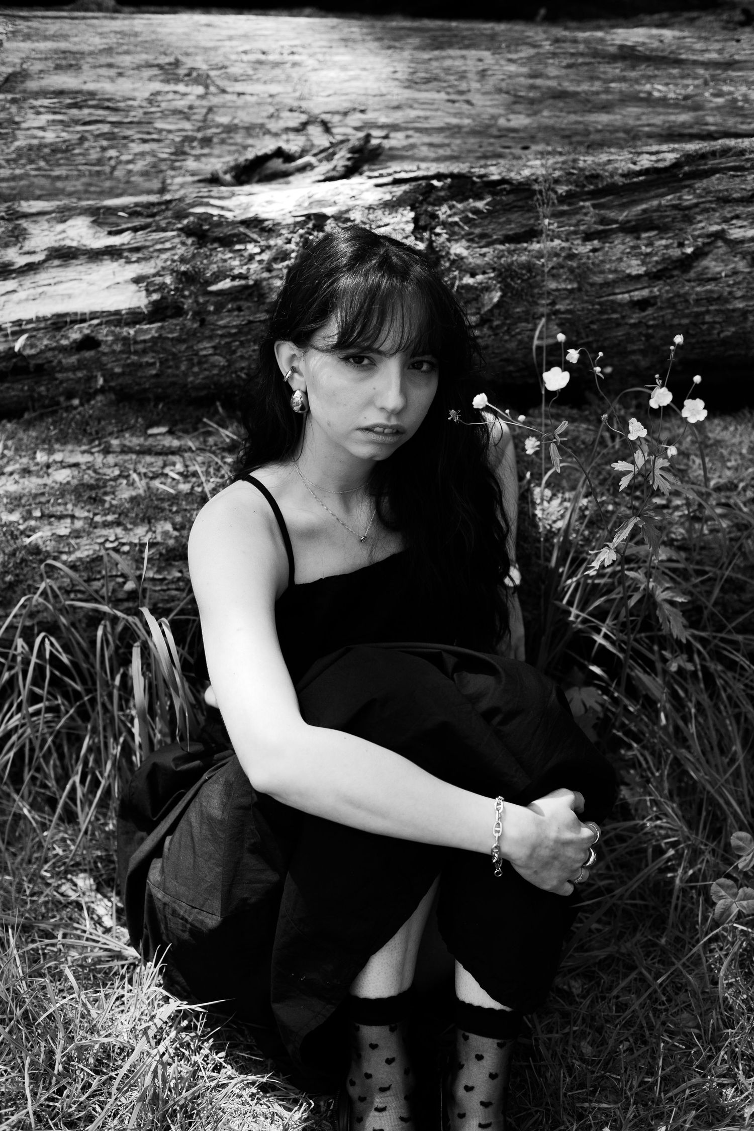 Black and white portrait of a young woman in black dress sitting near a fallen log outdoors.