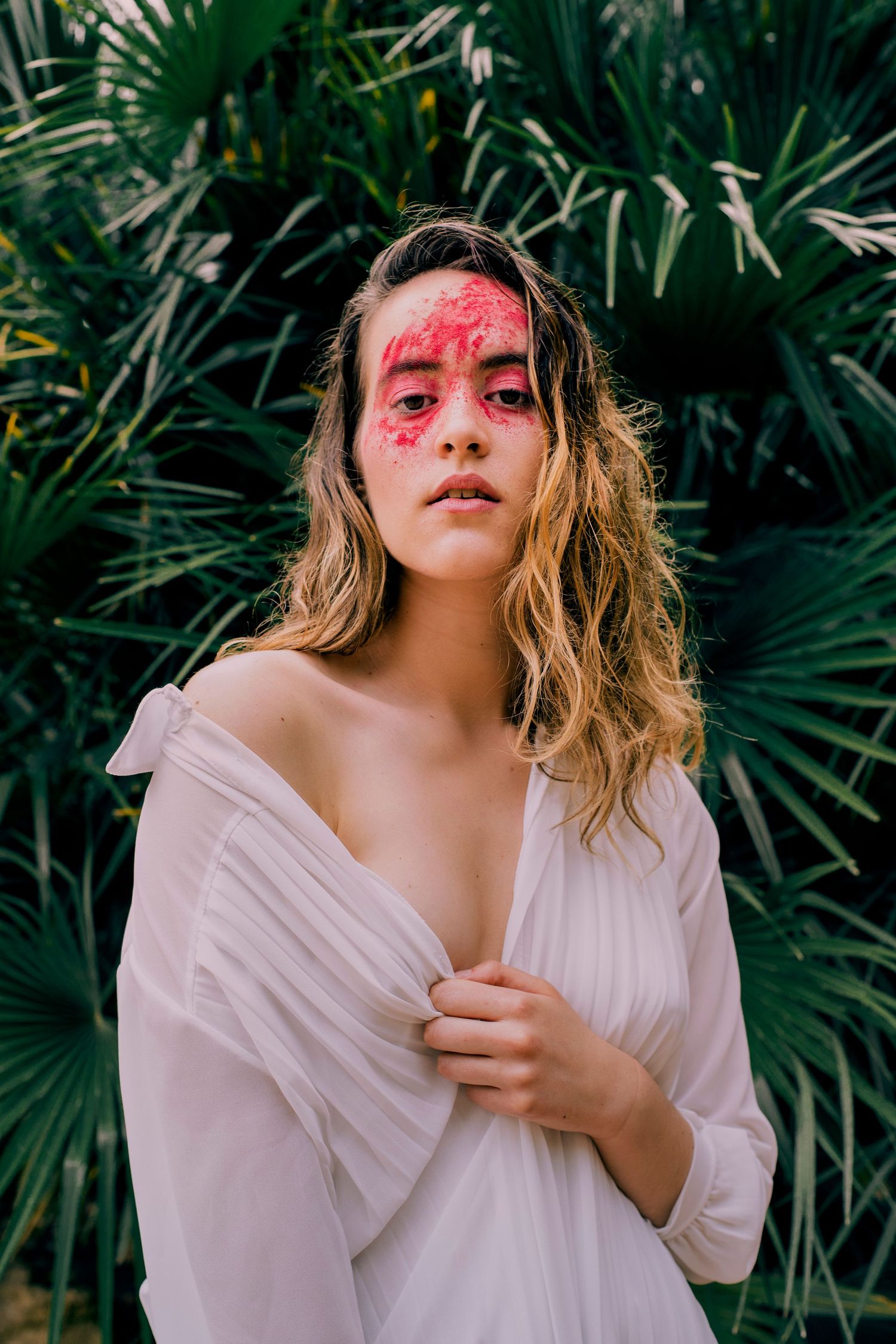 Woman with bold red face paint wearing white draped top before tropical palm plants.