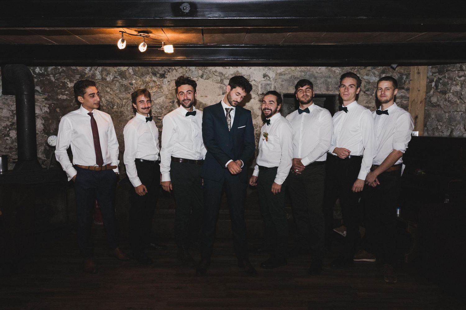 Groom in a navy suit posing with groomsmen in white shirts and bow ties against a stone wall.
