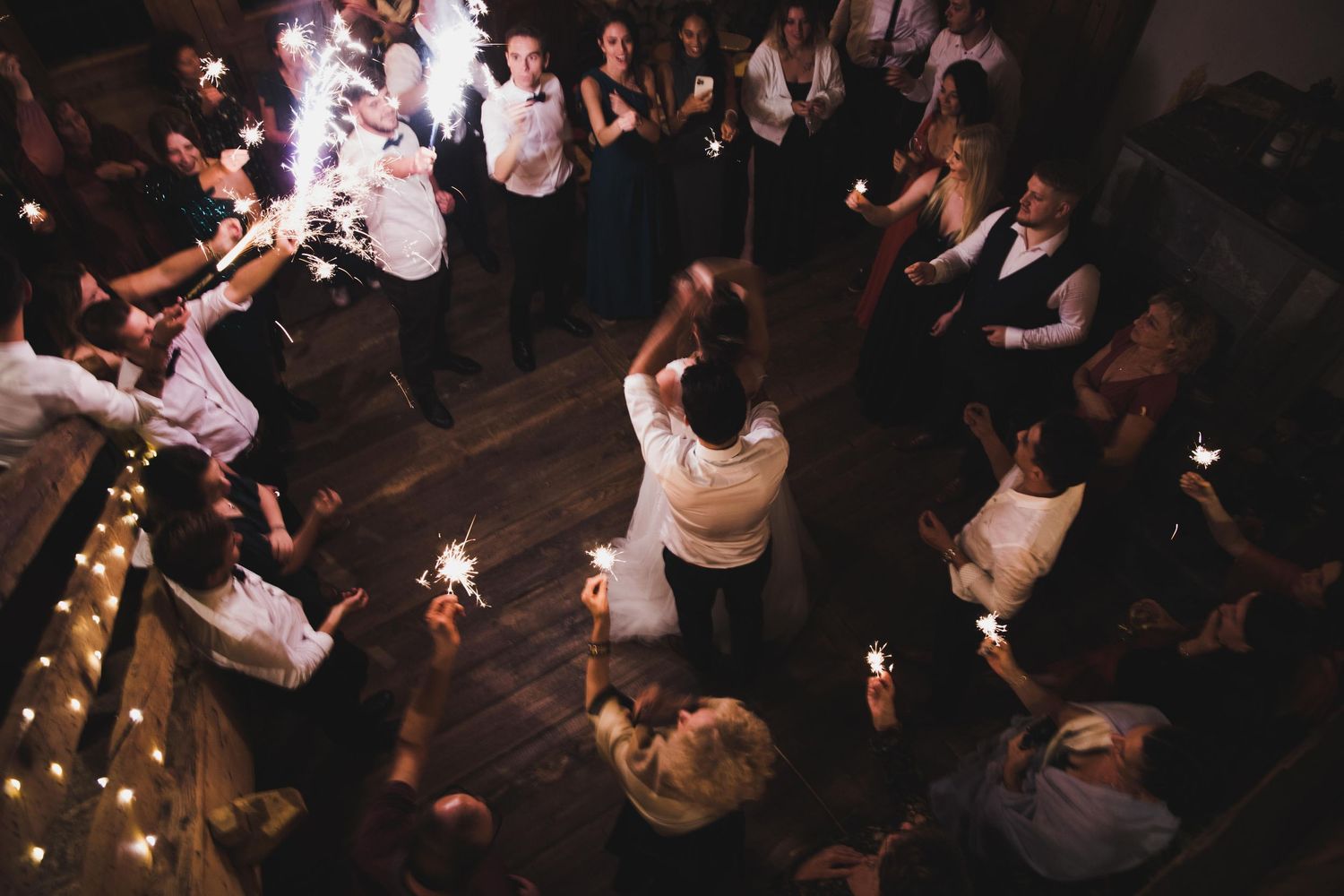 Wedding guests holding sparklers surround couple dancing at rustic barn reception.