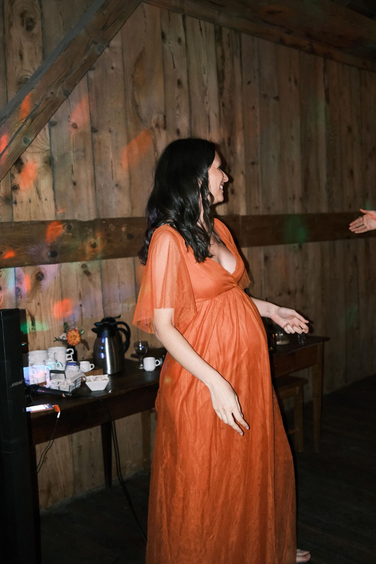 Pregnant woman in an elegant orange gown smiling at a rustic barn wedding reception.