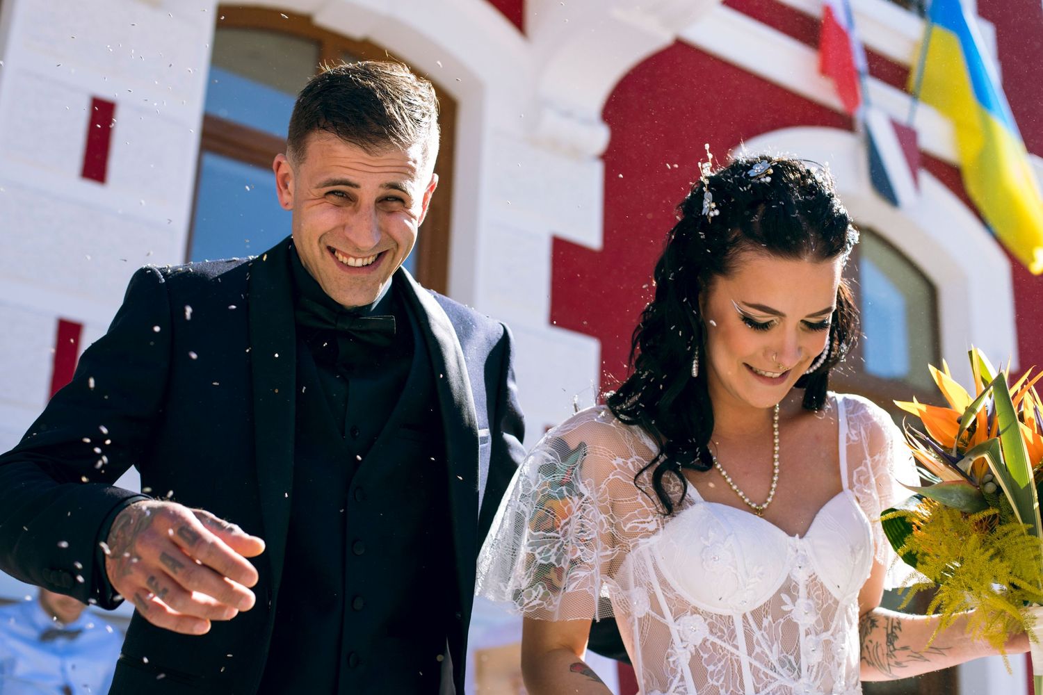 Smiling tattooed groom and dark-haired bride walk outside in sunlight with tropical wedding bouquet.
