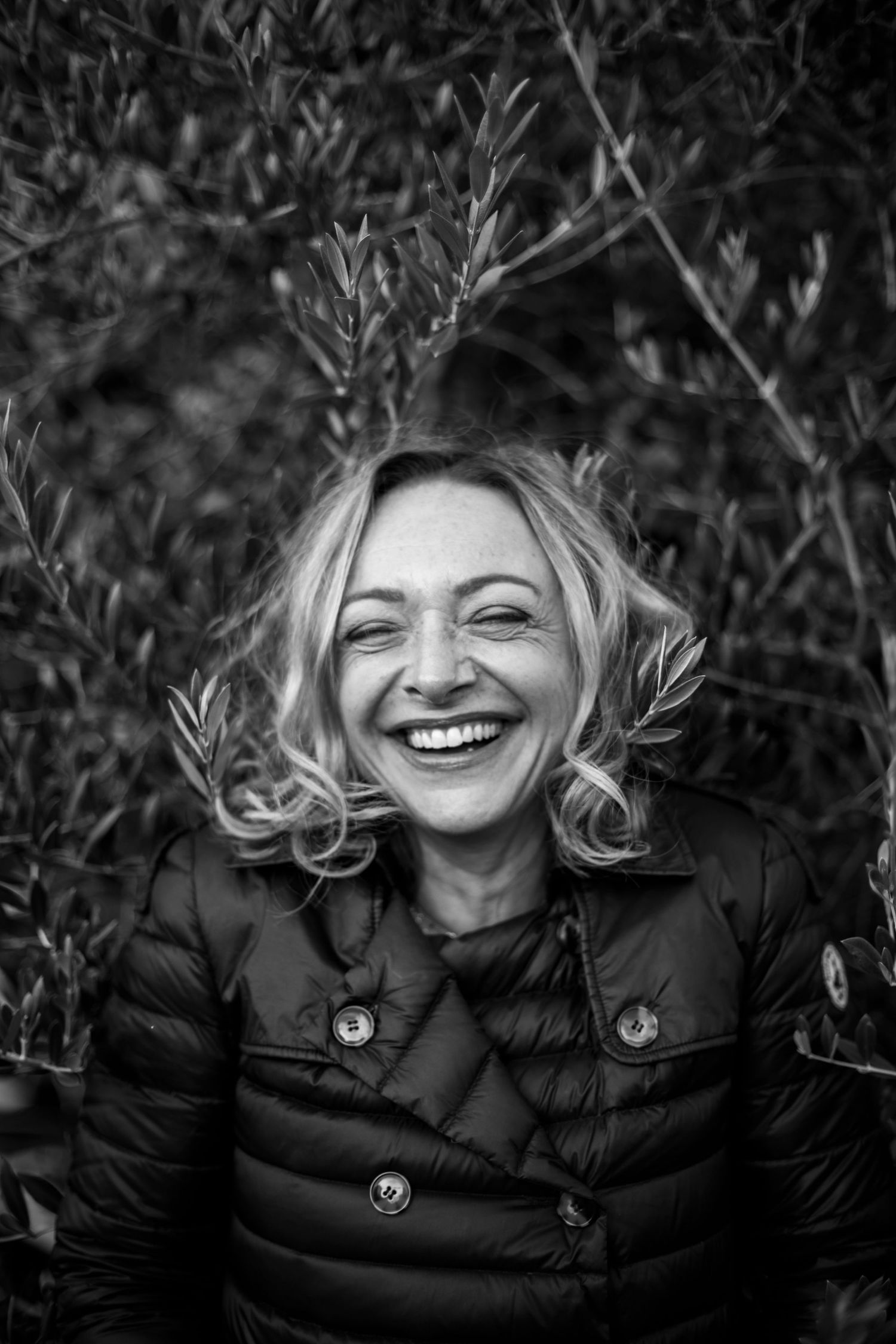 Black and white portrait of a laughing blonde woman in a dark jacket surrounded by leafy branches outdoors.