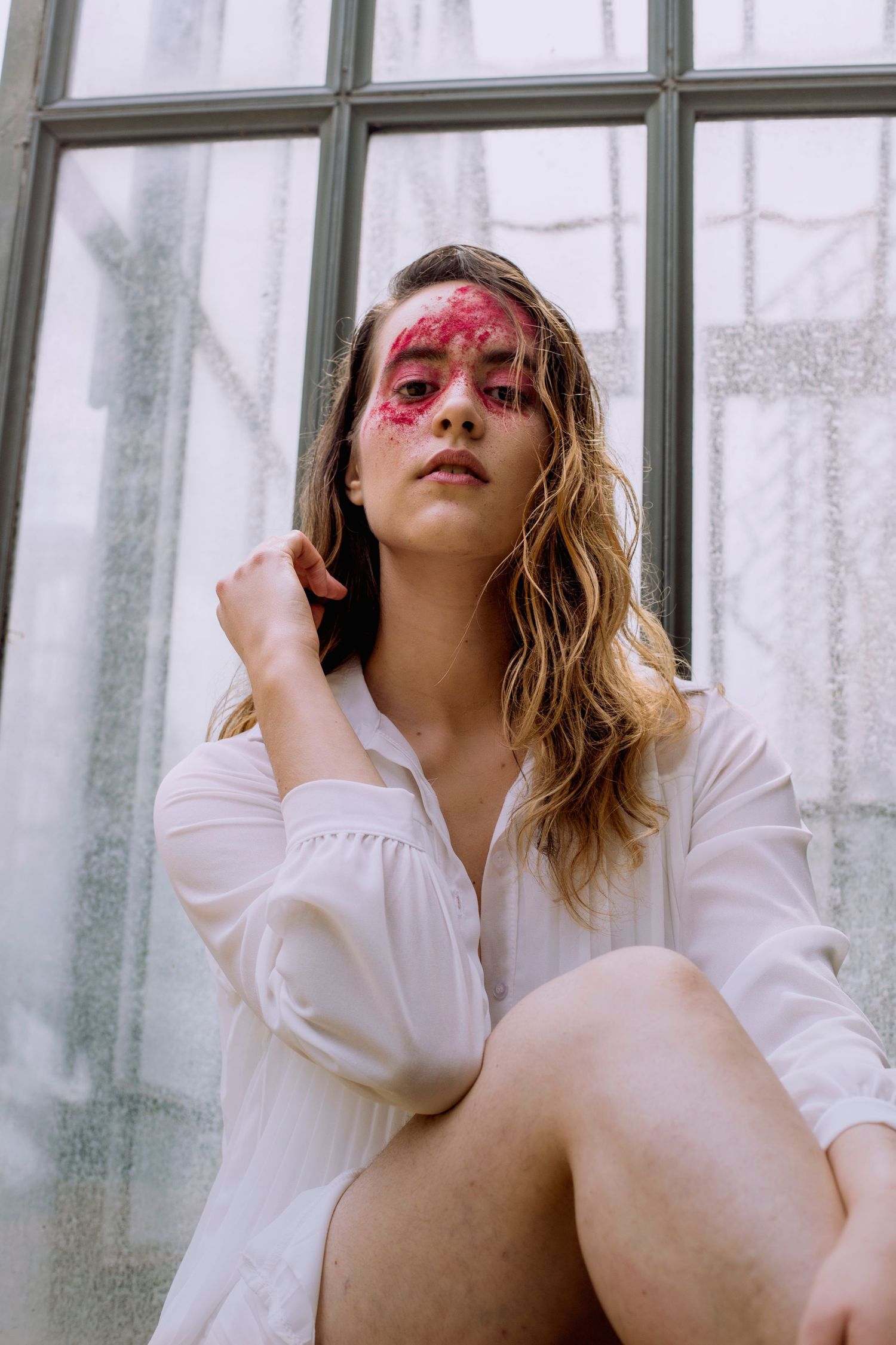 Woman with bold red face makeup sitting by large greenhouse windows.