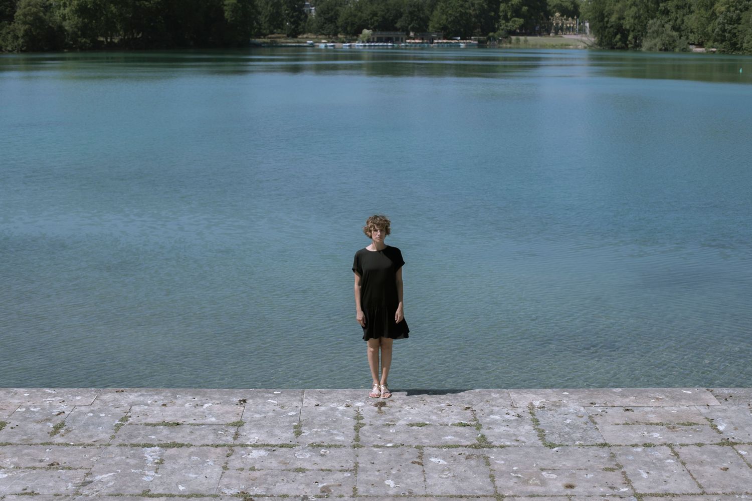 Woman in black dress standing on stone pavement beside a vast calm blue lake on a sunny day.