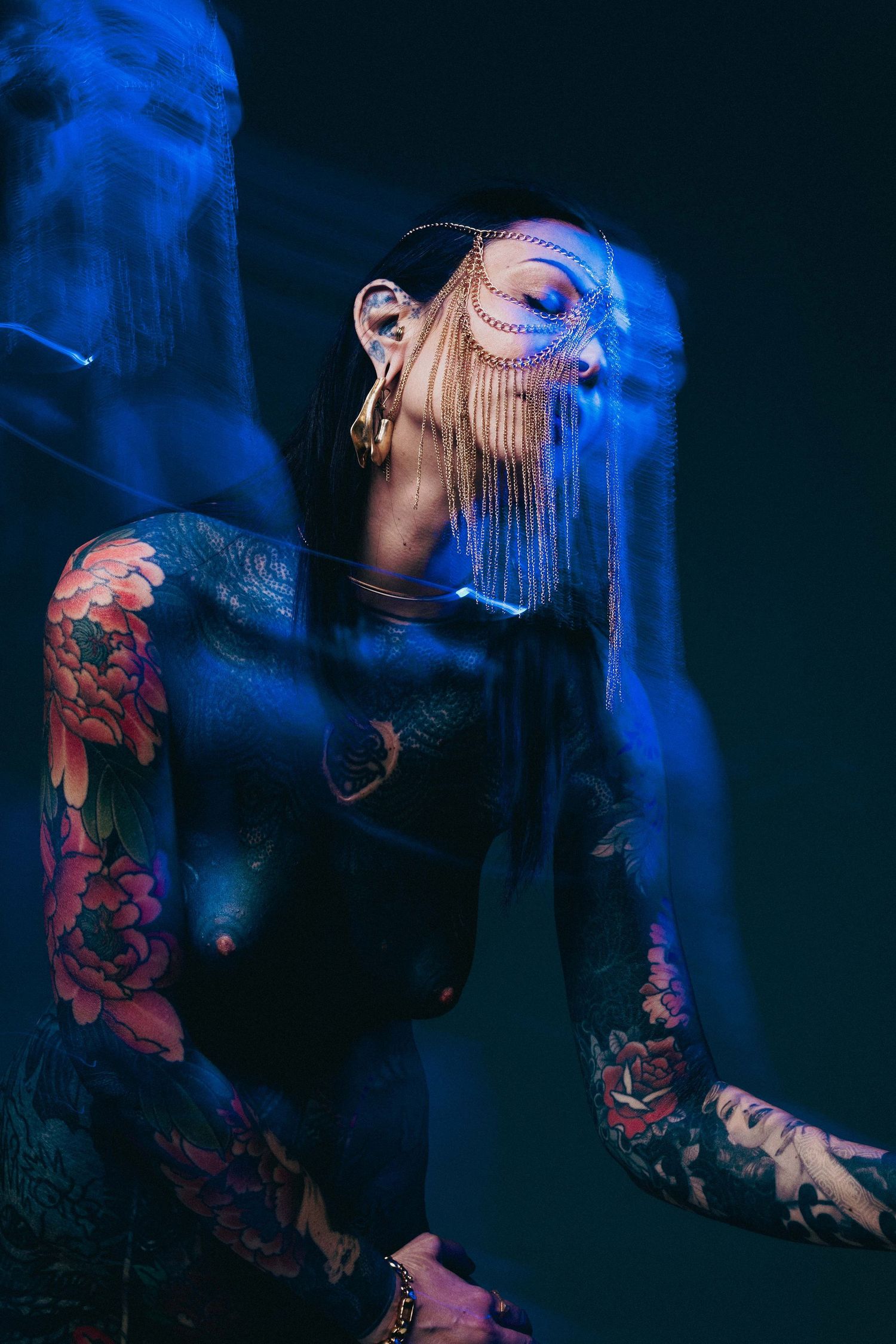 Heavily tattooed woman wearing ornate chain face mask under dramatic blue neon studio lighting.